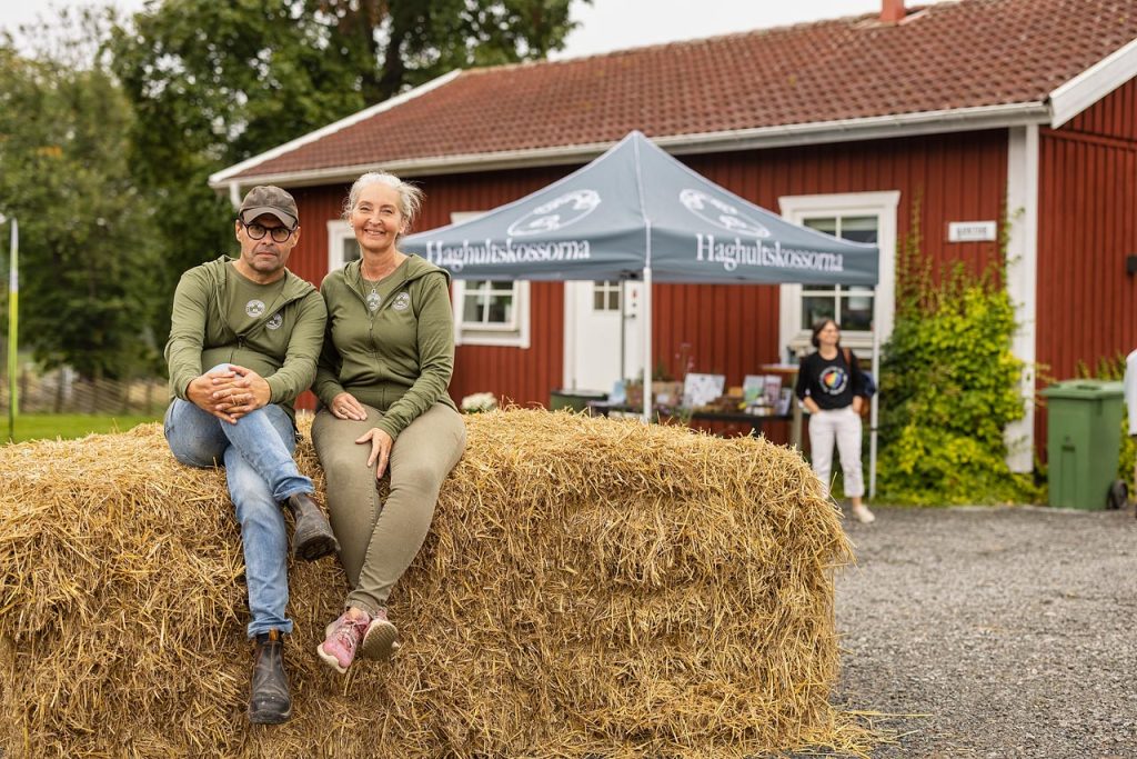 Två glada personer som sitter på halm framför ett rött hus